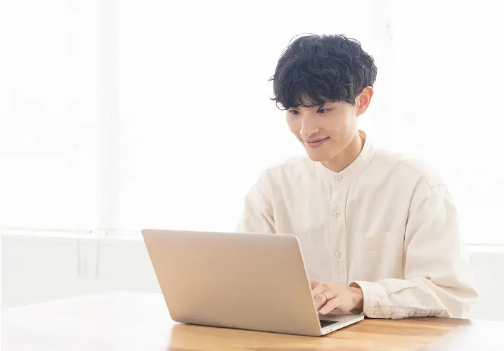 自宅でノートパソコンを操作し、笑顔で画面を見つめる若い男性の様子
