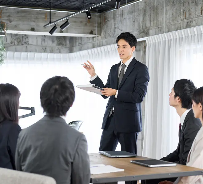 会議室でスーツ姿の男性が立ち上がり、資料を手に入社前研修を行う様子