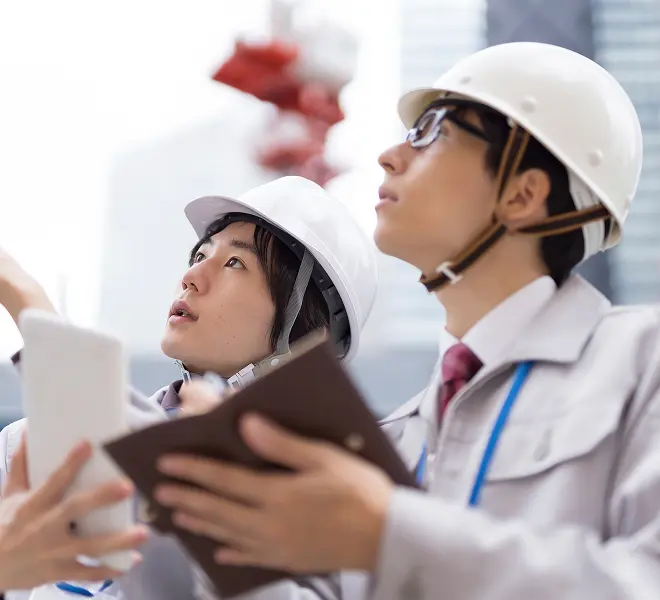 建設現場でヘルメットをかぶった技術者が図面を持ち、建物を見上げて確認している場面