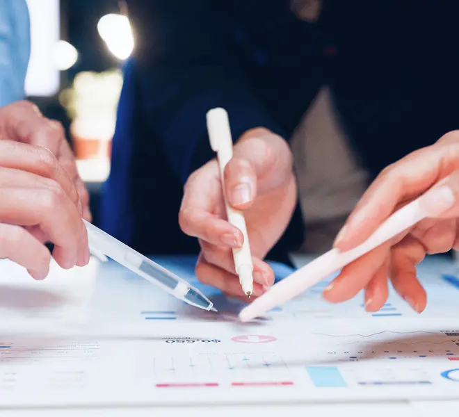 数人の手がペンを持ち、グラフや資料を指し示しながら議論している打ち合わせ風景