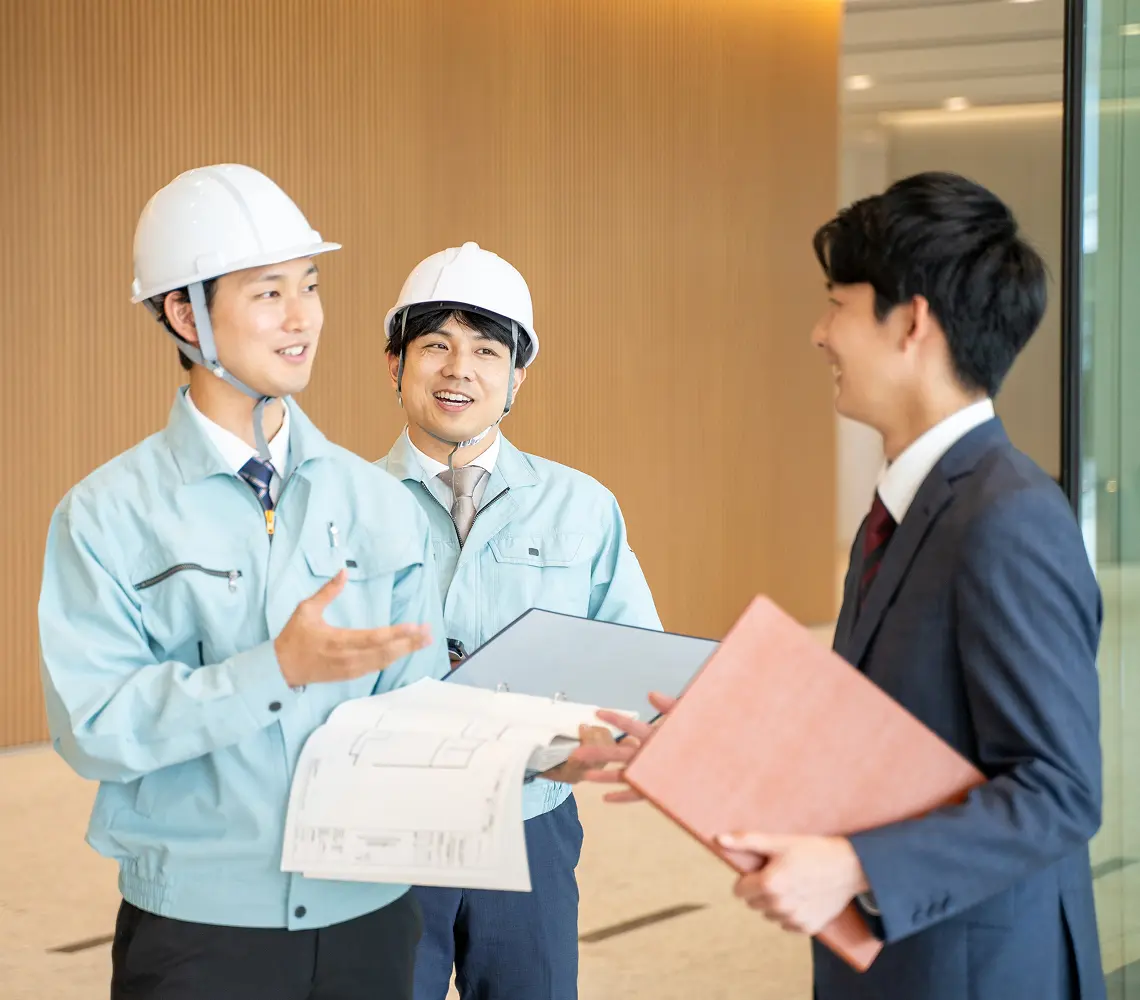 ヘルメットをかぶった作業員とスーツの人物が建築図面を見ながら相談している様子