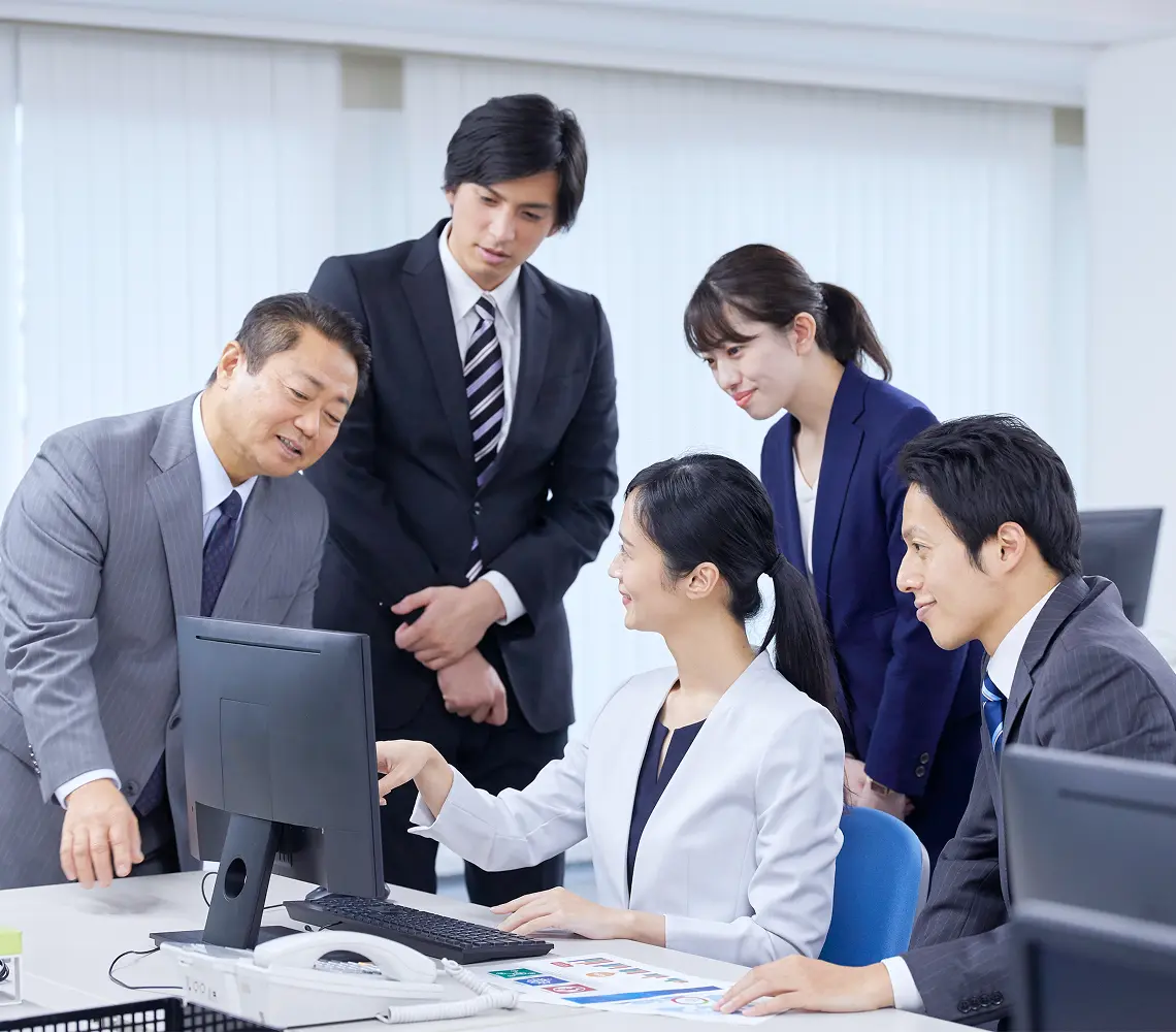 オフィスでスーツ姿の社員たちがパソコン画面を囲み、打ち合わせをする場面